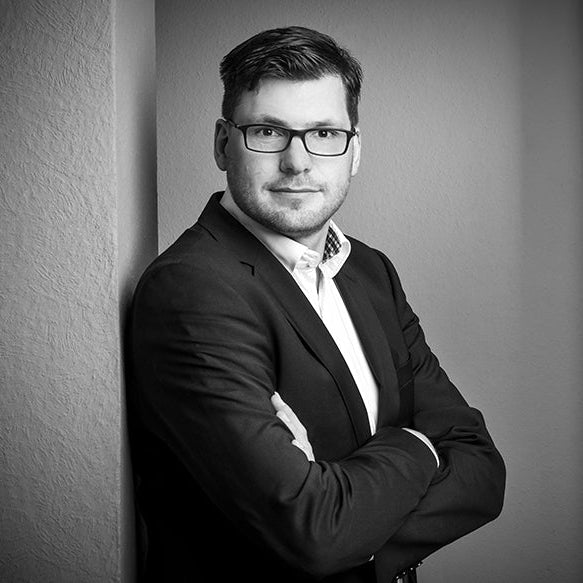 Mann im Anzug lehnt an Wand, Arme verschränkt, Brille tragend, schwarz-weiß Foto.