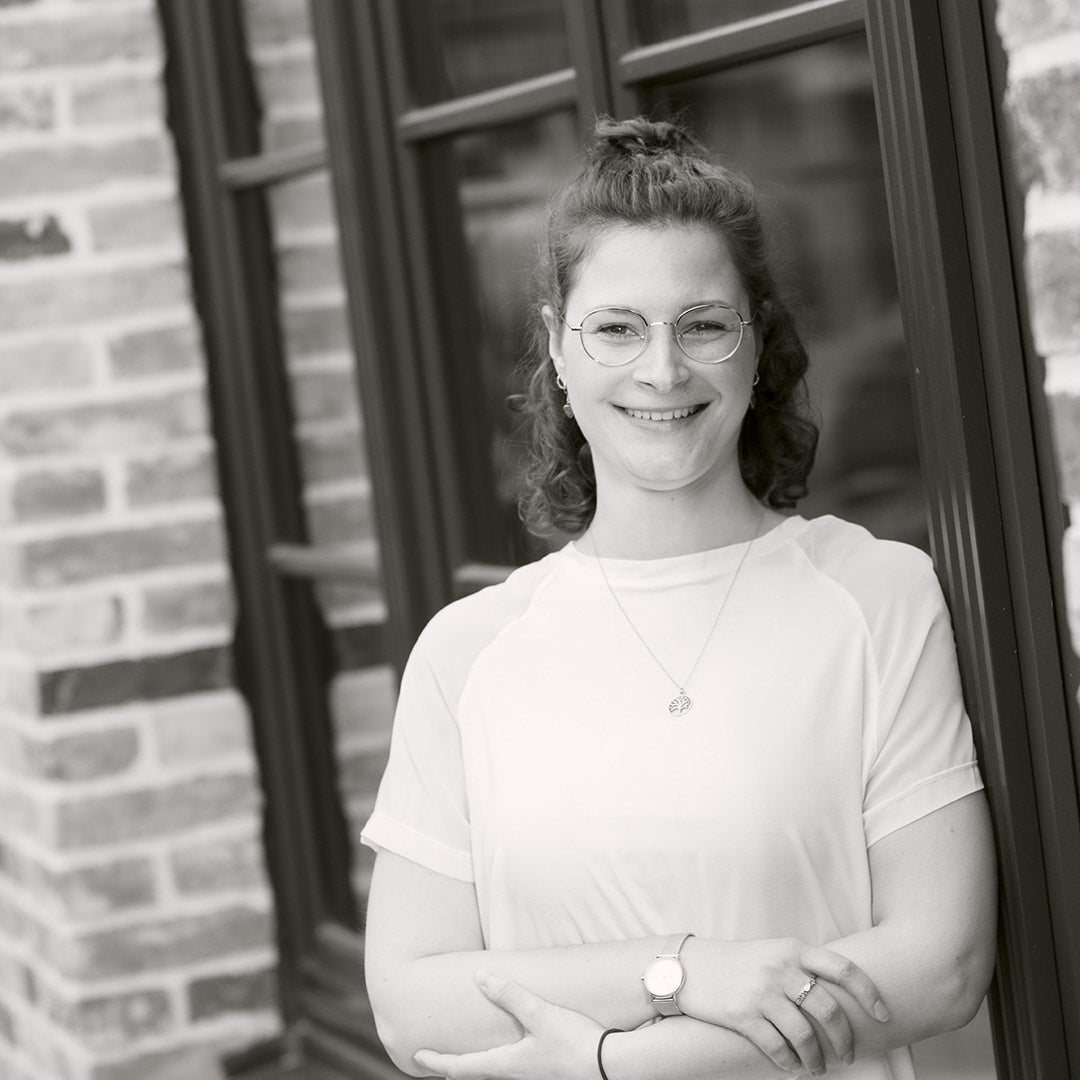 Frau mit Brille und lockigem Haar in weißem Oberteil steht lächelnd vor einer Wand.