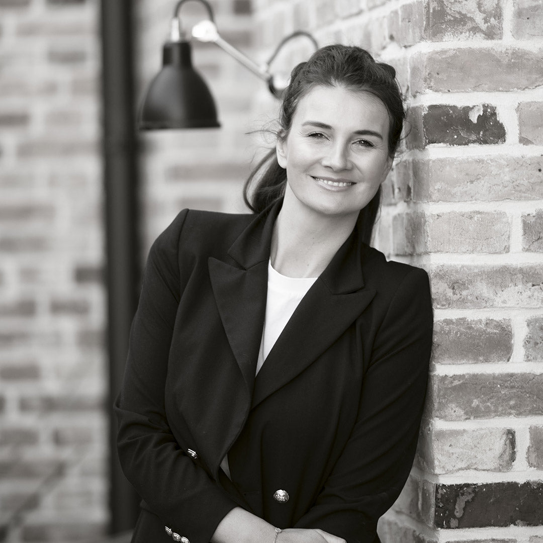 Frau im Blazer mit verschränkten Armen lehnt an Ziegelwand.