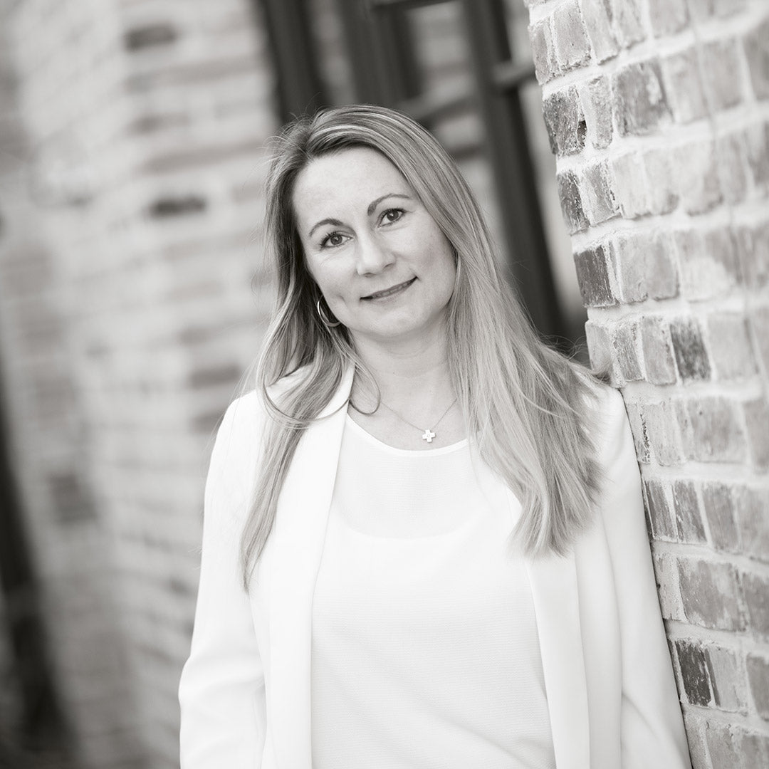 Frau mit langen Haaren und weißer Jacke lehnt an einer Ziegelwand.