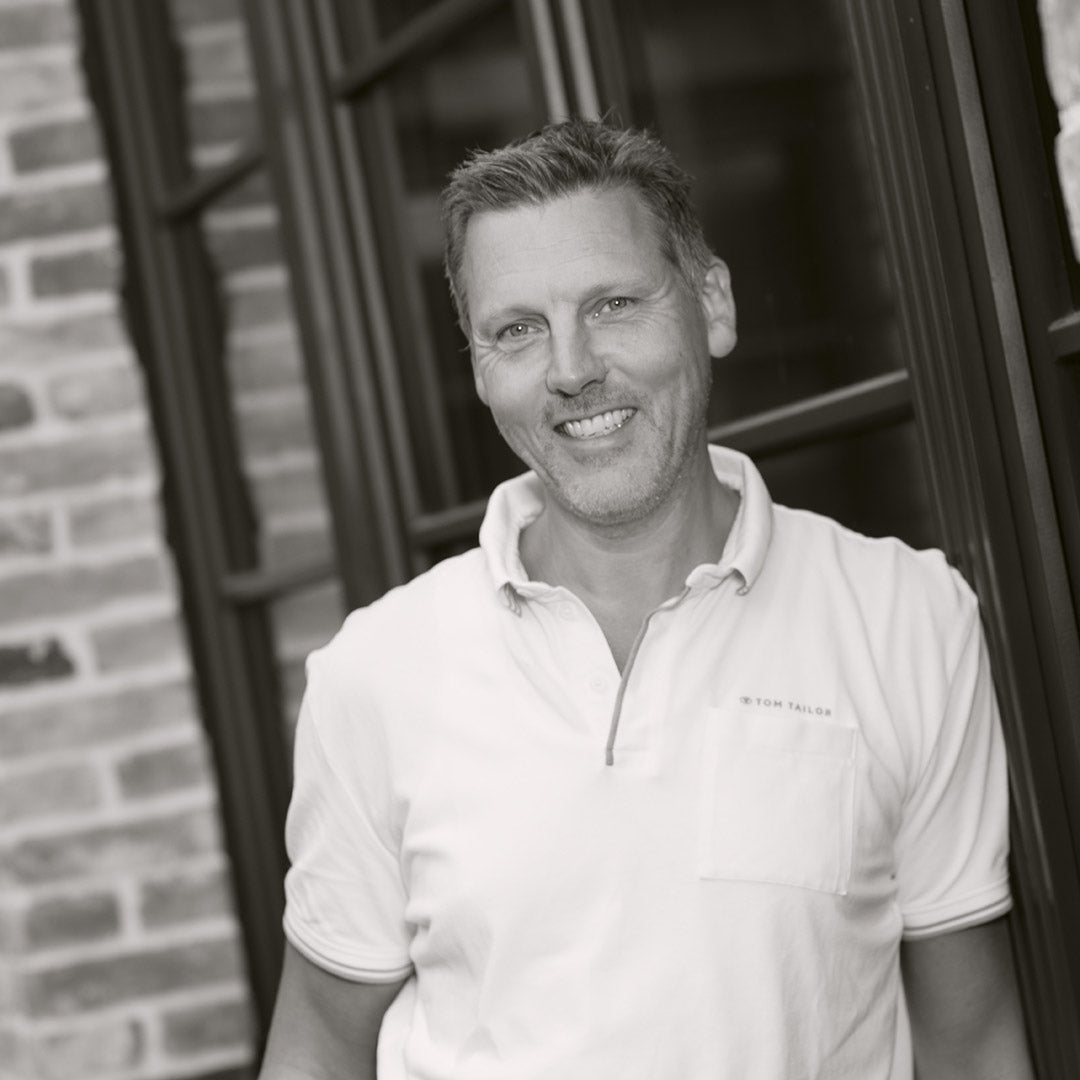 Mann mit kurzem Haar in einem weißen Poloshirt lächelt vor einer Backsteinfassade.