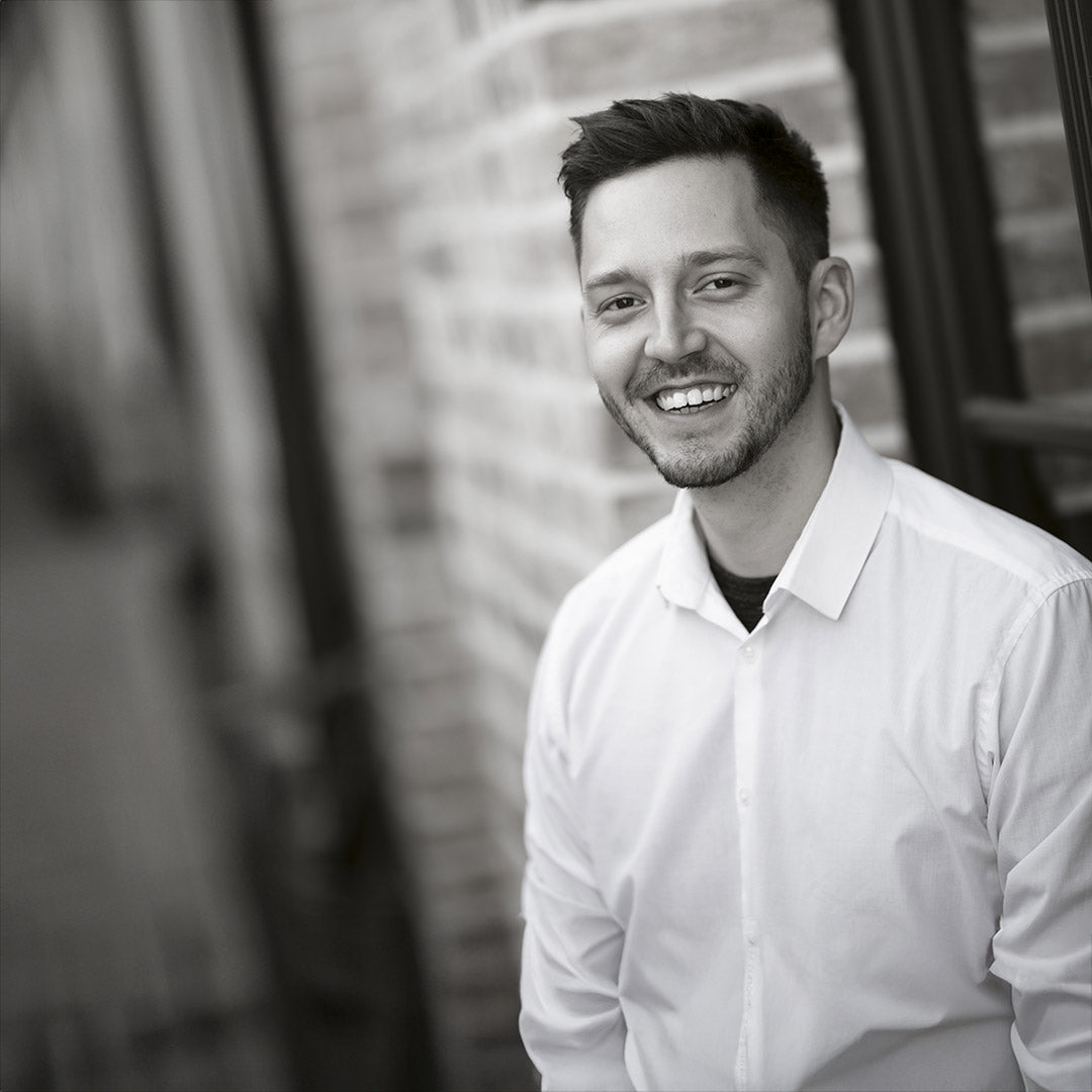 Lächelnder Mann in weißem Hemd lehnt an einer Ziegelwand.